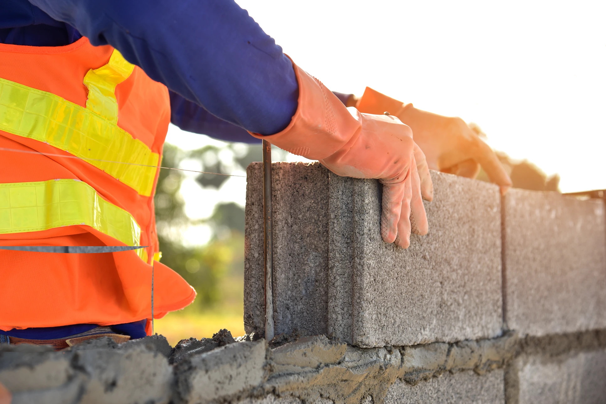 Ouvrier maçon en chemise à carreaux construisant un mur de briques rouges sur chantier extérieur, skyline urbain en arrière-plan flou au coucher du soleil